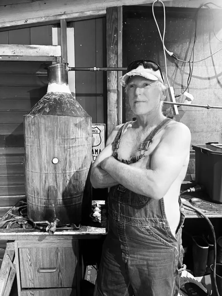 A man in overalls and a cap stands with arms crossed next to a large metal still in a rustic workshop.