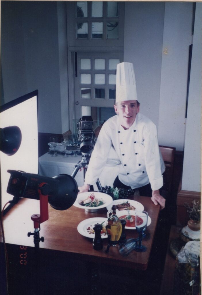 Chef in a white uniform and hat stands behind a table with prepared dishes, set up for a photoshoot with camera and lighting equipment.