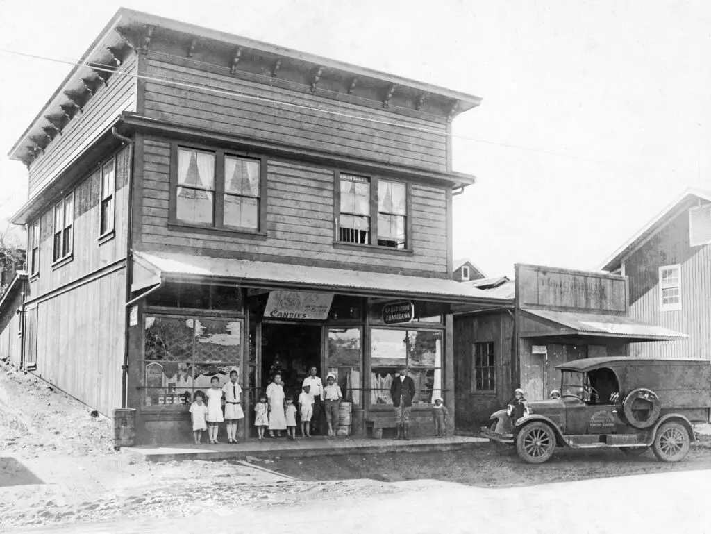 A group of people stands in front of a wooden building with a storefront, next to an early 20th-century car parked on the dirt road.