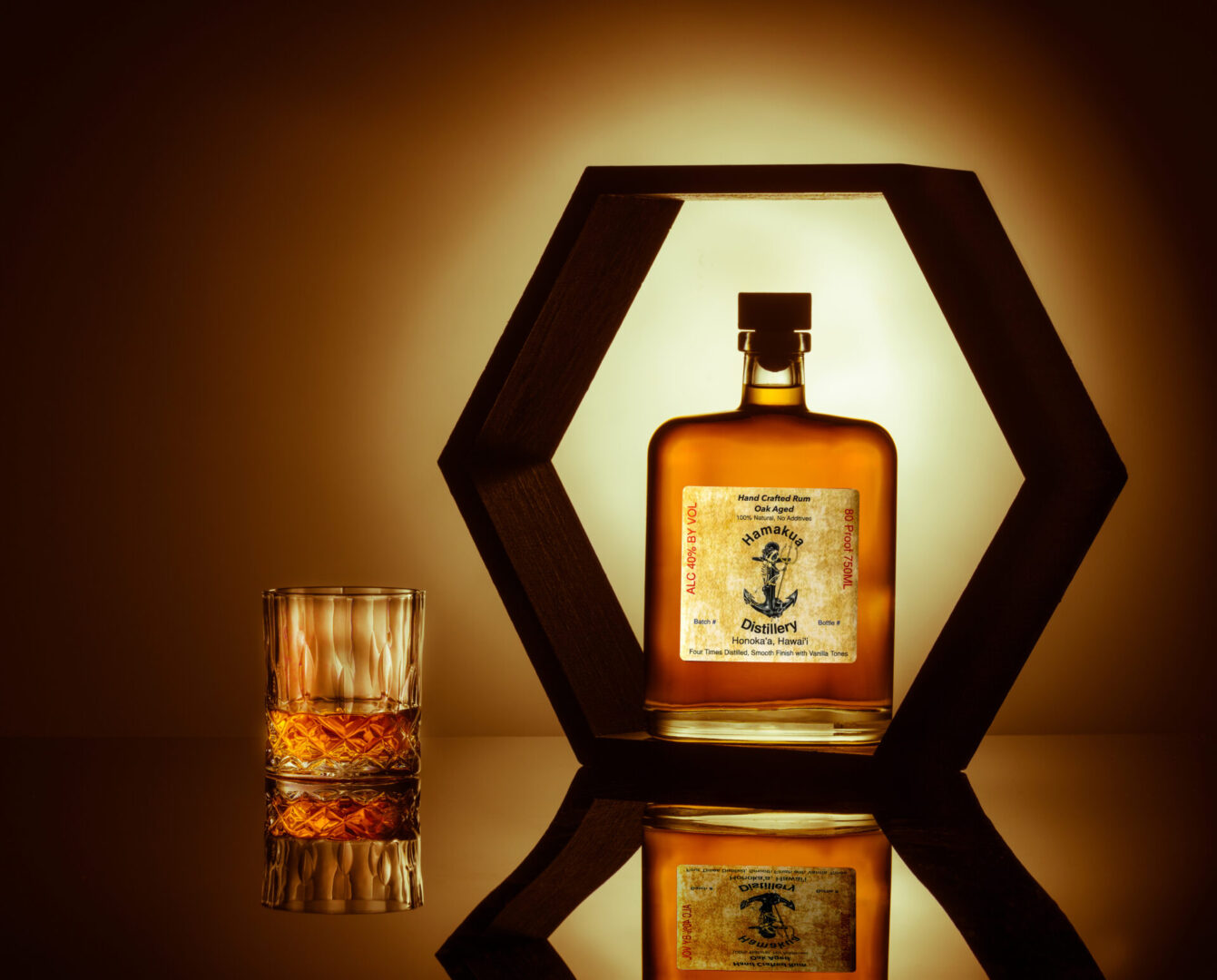 A hexagonal wooden frame holds a bottle of liquor with a nautical-themed label. A glass of whiskey sits beside it on a reflective surface, both illuminated by warm lighting.