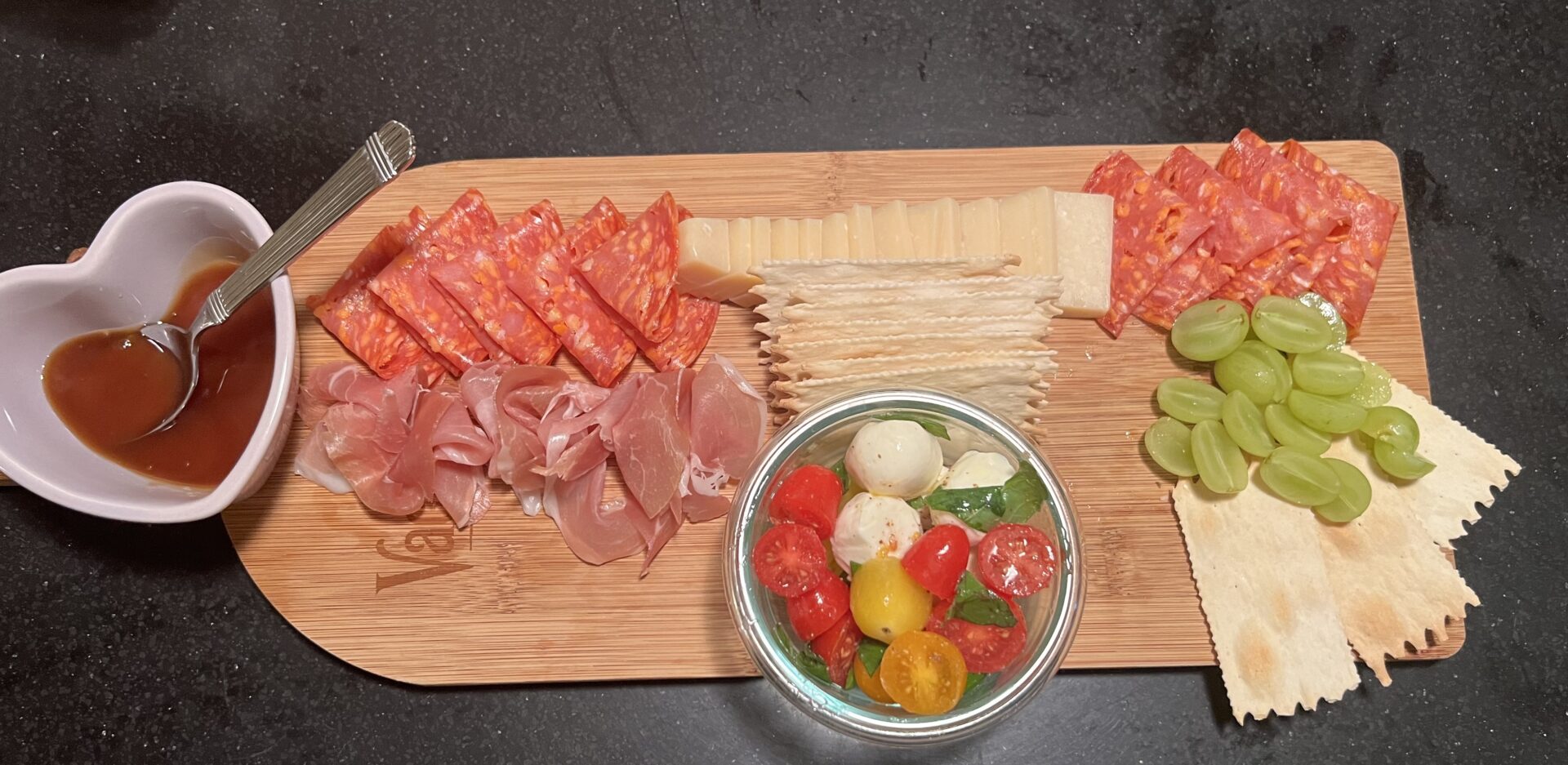 Charcuterie board with sliced meats, cheese, crackers, green grapes, caprese salad, and a heart-shaped dish of sauce on a wooden board.