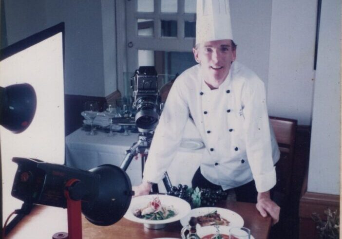 Chef in a white uniform and hat stands behind a table with prepared dishes, set up for a photoshoot with camera and lighting equipment.