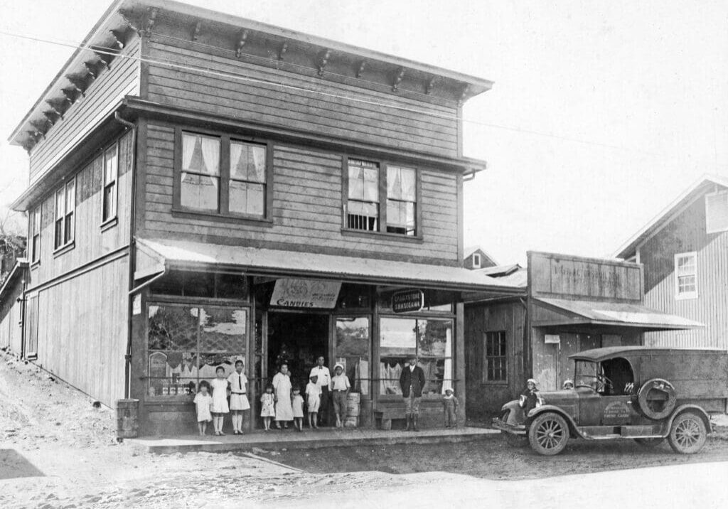 A group of people stands in front of a wooden building with a storefront, next to an early 20th-century car parked on the dirt road.