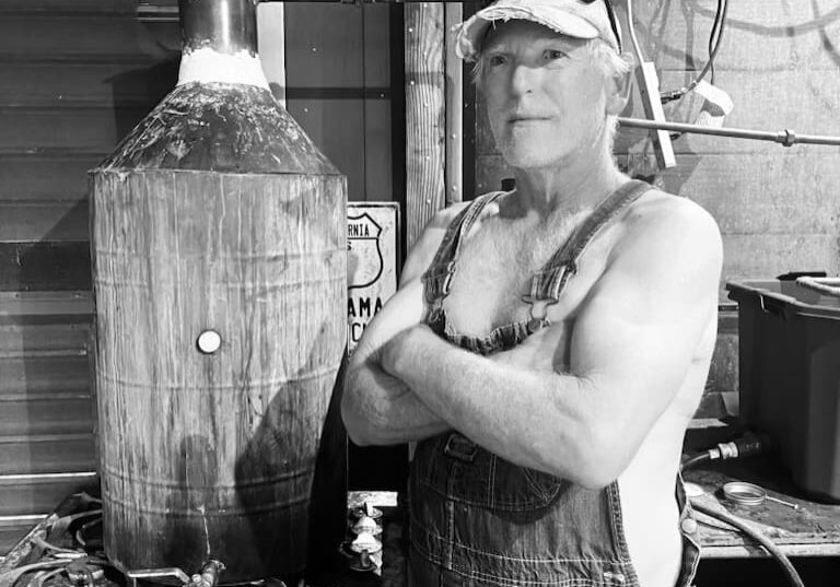 A man in overalls and a cap stands with arms crossed next to a large metal still in a rustic workshop.